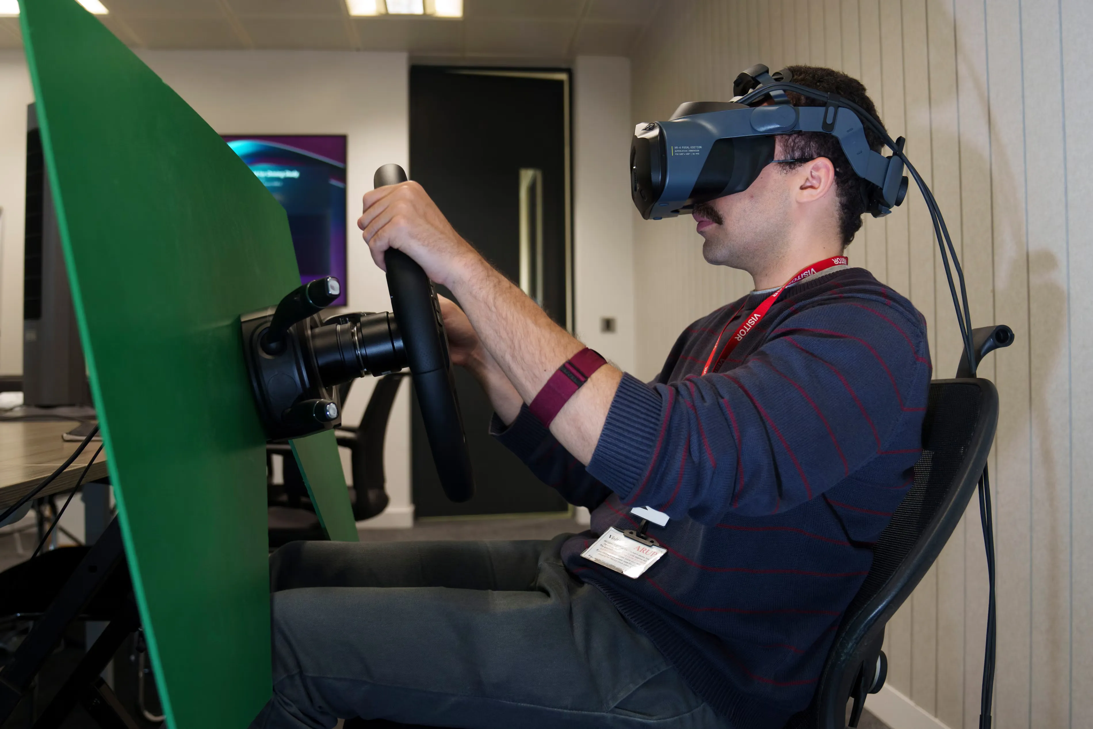 A person sitting wearing a VR headset with a steering wheel in front of a green panel.